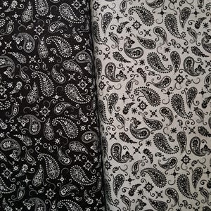 May include: Two rolls of fabric featuring a repeating paisley pattern. The left roll is black with a white paisley print, while the right roll is white with a black paisley print. Both fabrics display intricate teardrop shapes and floral motifs.