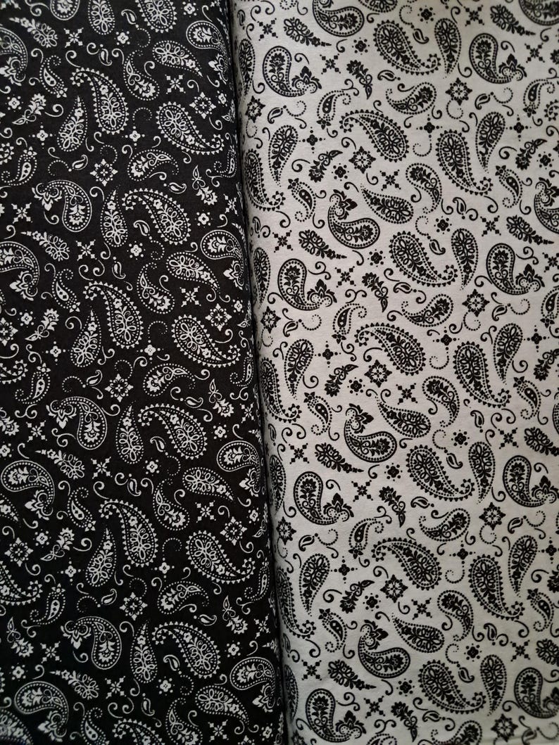 May include: Two rolls of fabric featuring a repeating paisley pattern. The left roll is black with a white paisley print, while the right roll is white with a black paisley print. Both fabrics display intricate teardrop shapes and floral motifs.