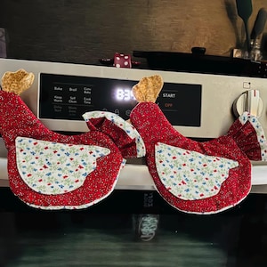 May include: Two fabric oven mitts shaped like chickens, featuring a red body with a small white floral pattern, a white wing with a blue and red floral print, and a gold-toned textured comb on top. The mitts are displayed hanging on a silver oven.