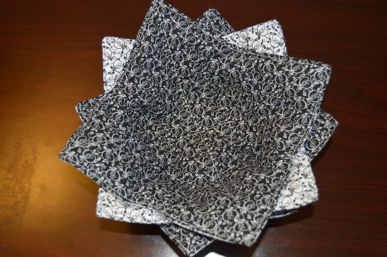May include: A fabric bowl cozy featuring a black and white floral scroll pattern. The square-shaped holder has eight pointed corners and is constructed with quilted fabric, showcasing a contrasting black interior and a white exterior design.