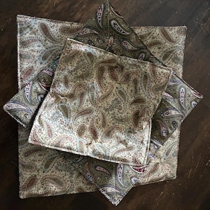 May include: A set of three square fabric bowl cozies featuring a repeating paisley pattern in shades of olive green, cream, tan, and deep red. The quilted fabric items are stacked, showing a consistent print design with stitched edges.