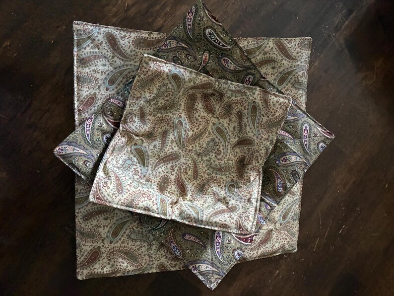 May include: A set of three square fabric bowl cozies featuring a repeating paisley pattern in shades of olive green, cream, tan, and deep red. The quilted fabric items are stacked, showing a consistent print design with stitched edges.