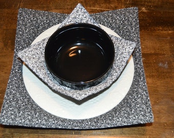 May include: A square fabric bowl cozy featuring a black and white floral pattern. The cozy holds a round white plate and a small, smooth, black ceramic bowl centered within the fabric structure, creating a layered, functional kitchen accessory.