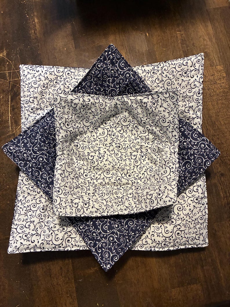 May include: Two square fabric bowl cozies featuring a navy blue and white swirling vine pattern. The larger cozy has a white background with navy vines, while the smaller nested cozy has a navy background with white vines, both quilted.