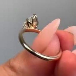 May include: A gold ring with a marquise-cut diamond center stone and a delicate, floral-inspired setting. The ring's band is smooth and polished, reflecting light. The diamond is clear and sparkling, set in a prong setting.