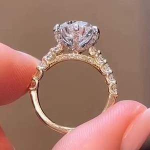 Può includere: Un anello di fidanzamento in oro con un grande diamante rotondo e chiaro al centro e diamanti più piccoli incastonati a pavé attorno alla fascia.