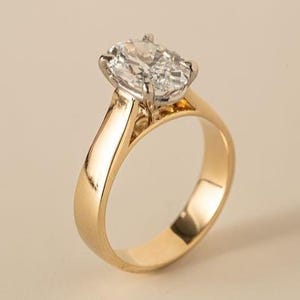 May include: A gold engagement ring with a large, oval-cut diamond. The ring features a classic, polished gold band and a four-prong setting that securely holds the sparkling gemstone. The ring is set against a neutral background.