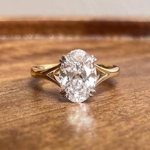 May include: A gold engagement ring with a large, oval-cut diamond. The diamond is set in a four-prong setting, and the band has a split shank design. The ring is displayed on a wooden surface.
