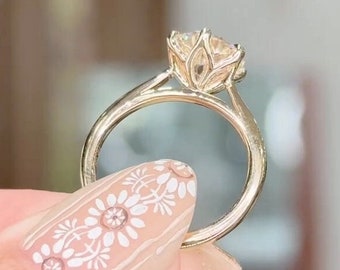 Puede incluir: Un anillo de compromiso de oro con un gran diamante redondo y claro engastado en un engaste con forma de flor.