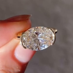 Puede incluir: Un anillo de oro con un gran diamante de talla ovalada. El diamante está engastado en un bisel y la banda es de oro pulido. El anillo se sostiene sobre un fondo neutro.