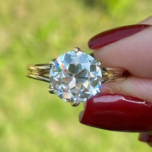 Puede incluir: Un anillo de oro con una gema grande, clara y facetada. El anillo tiene un diseño clásico con una caña dividida y un engaste de garras que sostiene la piedra brillante. El fondo es de un verde suave.