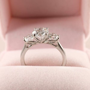 May include: A three-stone engagement ring with a large, round center diamond and two smaller side stones. The ring is set in a silver band and presented in a pink velvet ring box. The diamonds sparkle brightly.