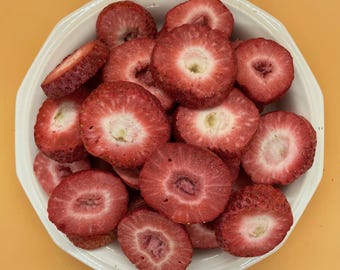May include: A white, octagonal bowl filled with sliced strawberries. The strawberries are a vibrant red with white centers, and the bowl sits on an orange surface. The image is a top-down view of the fruit.