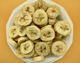 May include: A white ceramic bowl filled with dried banana slices. The banana slices are circular, with a light yellow color and a slightly darker center. The bowl is set against an orange background.