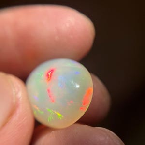 May include: A smooth, rounded, translucent white opal gemstone featuring a vibrant play-of-color with distinct flashes of bright red, orange, and green light across its polished surface, showcasing a natural iridescent quality throughout the stone.