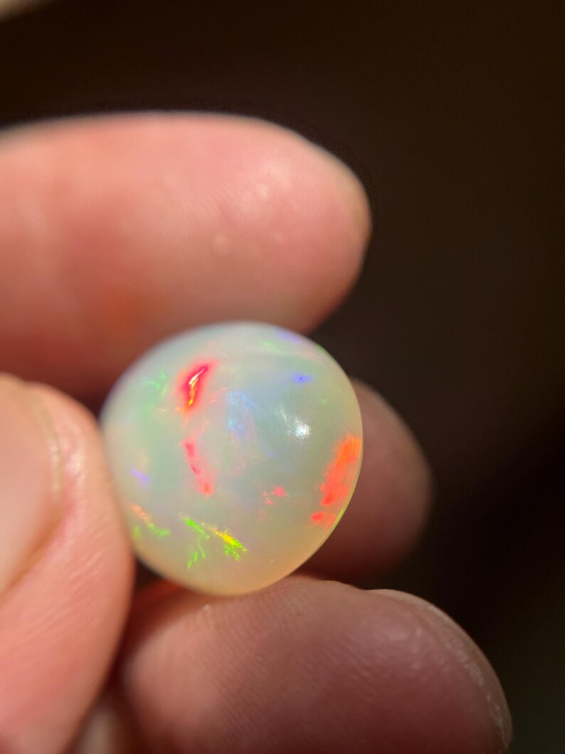 May include: A smooth, rounded, translucent white opal gemstone featuring a vibrant play-of-color with distinct flashes of bright red, orange, and green light across its polished surface, showcasing a natural iridescent quality throughout the stone.