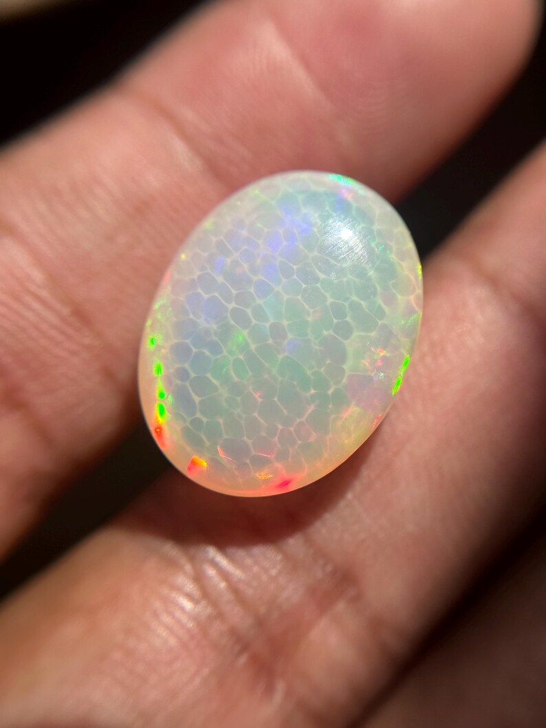 May include: An oval-shaped cabochon opal gemstone featuring a translucent white base with a distinct honeycomb pattern. The stone displays vibrant flashes of green, blue, and orange play-of-color throughout its polished, smooth, and rounded surface.