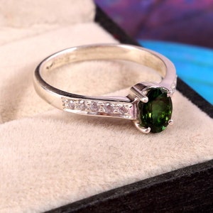 May include: A silver ring featuring a vibrant green oval gemstone as the centerpiece. The band is adorned with small, clear accent stones. The ring is set against a neutral background, highlighting its elegant design.