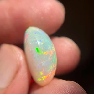 May include: A smooth, oval-shaped white opal gemstone featuring a translucent body with vibrant flashes of neon green, orange, and blue color play throughout the surface. The stone has a polished, rounded cabochon cut that reflects light.