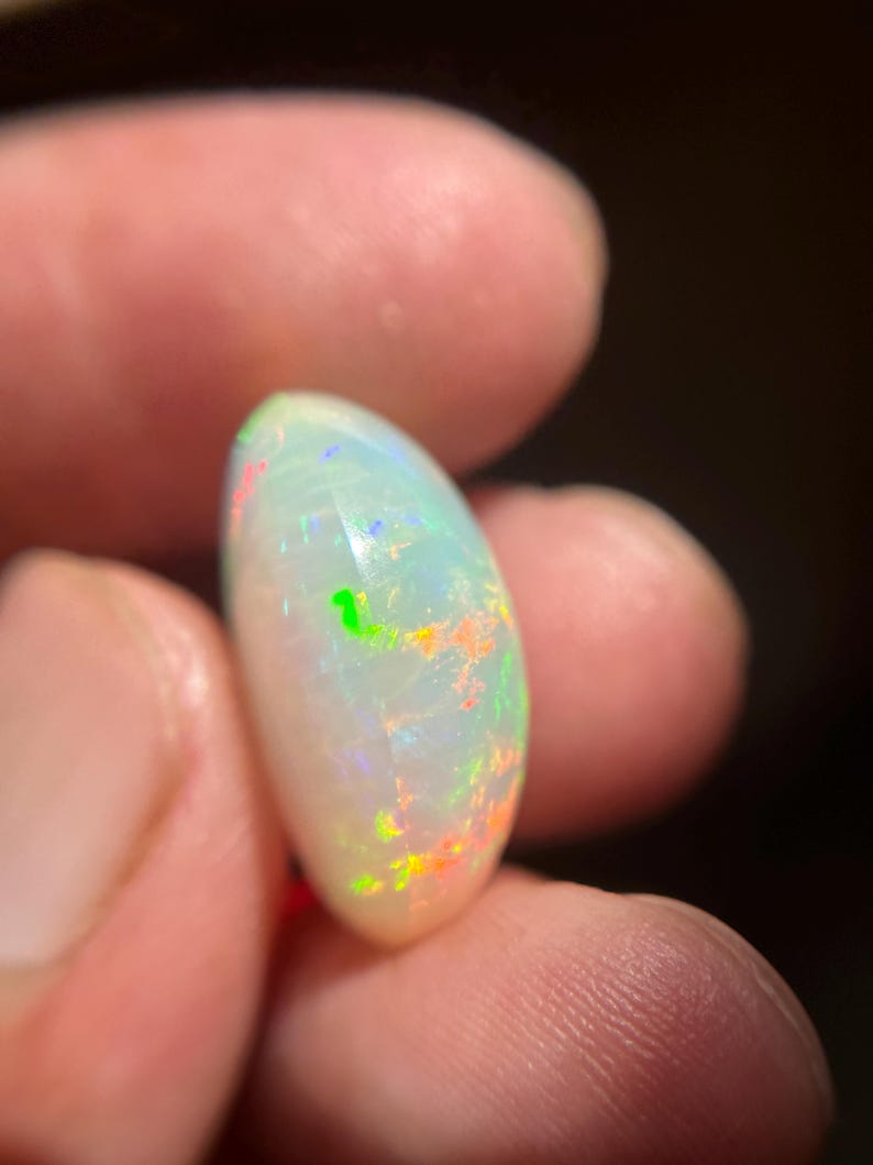 May include: A smooth, oval-shaped white opal gemstone featuring a translucent body with vibrant flashes of neon green, orange, and blue color play throughout the surface. The stone has a polished, rounded cabochon cut that reflects light.
