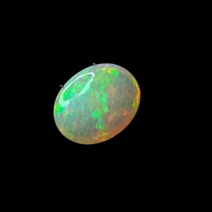 May include: An oval-shaped opal gemstone featuring a translucent, milky white base with vibrant flashes of neon green and subtle orange play-of-color throughout its surface. The stone displays a smooth, polished cabochon cut with light reflections.
