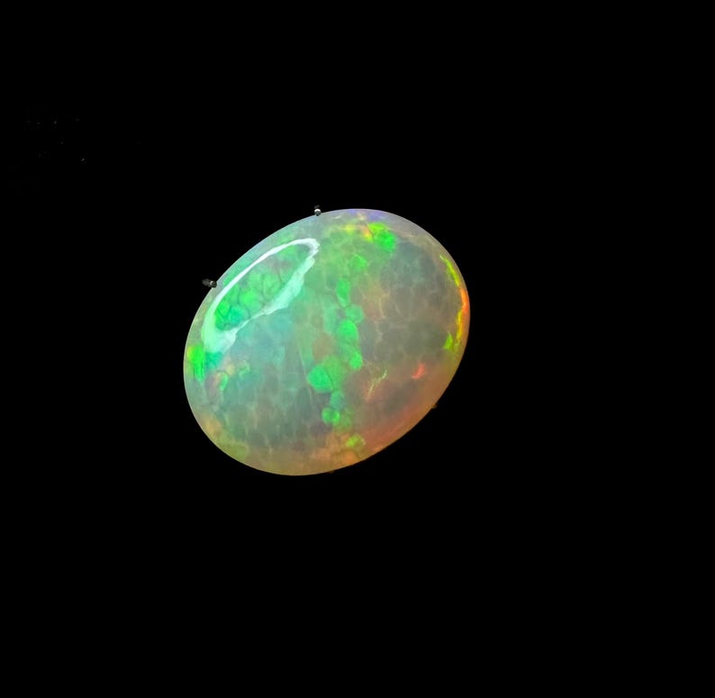 May include: An oval-shaped opal gemstone featuring a translucent, milky white base with vibrant flashes of neon green and subtle orange play-of-color throughout its surface. The stone displays a smooth, polished cabochon cut with light reflections.