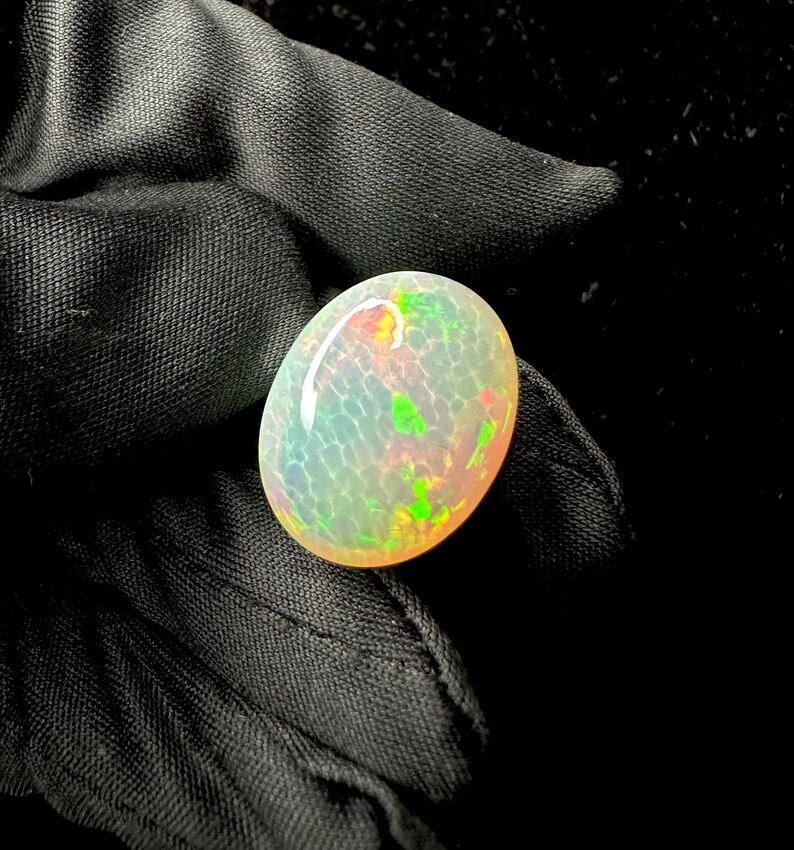 May include: An oval-shaped cabochon opal gemstone featuring a translucent white base with a distinct honeycomb pattern. The stone displays vibrant flashes of green, orange, and red play-of-color across its smooth, polished, and domed surface.