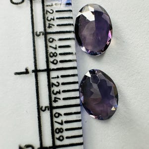 May include: Two oval-shaped, faceted purple gemstones positioned next to a clear plastic ruler. The ruler shows the stones measure approximately 0.5 inches in length. The gemstones have a translucent appearance with visible light reflections.