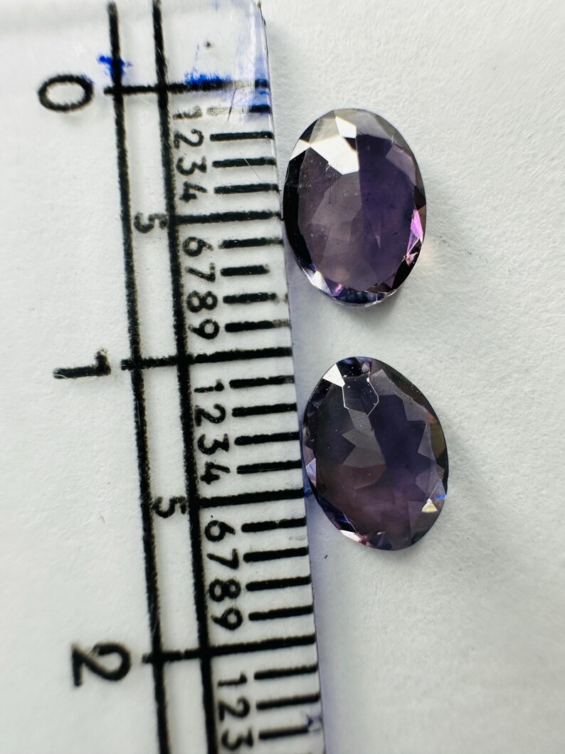 May include: Two oval-shaped, faceted purple gemstones positioned next to a clear plastic ruler. The ruler shows the stones measure approximately 0.5 inches in length. The gemstones have a translucent appearance with visible light reflections.