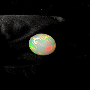 May include: An oval-shaped cabochon opal gemstone featuring a milky white base color with vibrant flashes of red, orange, yellow, and green play-of-color throughout the surface, displayed against a dark, textured fabric background.