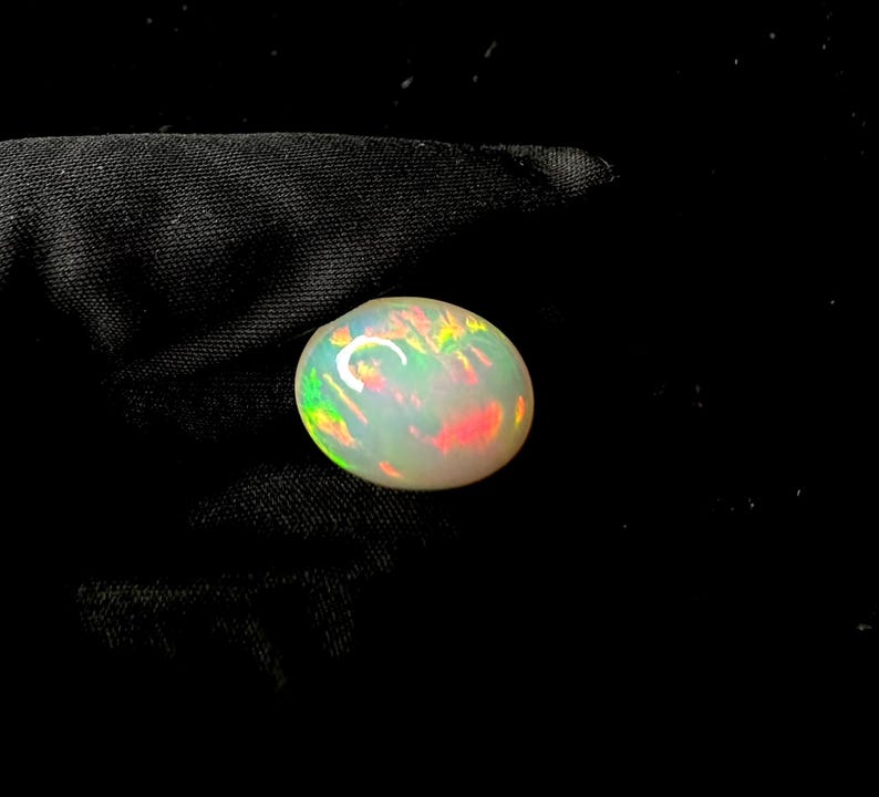 May include: An oval-shaped cabochon opal gemstone featuring a milky white base color with vibrant flashes of red, orange, yellow, and green play-of-color throughout the surface, displayed against a dark, textured fabric background.