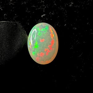 May include: An oval-shaped cabochon opal gemstone featuring a translucent, milky white base with vibrant, scattered flashes of neon green and bright orange color play across its smooth, polished, and rounded surface against a dark background.
