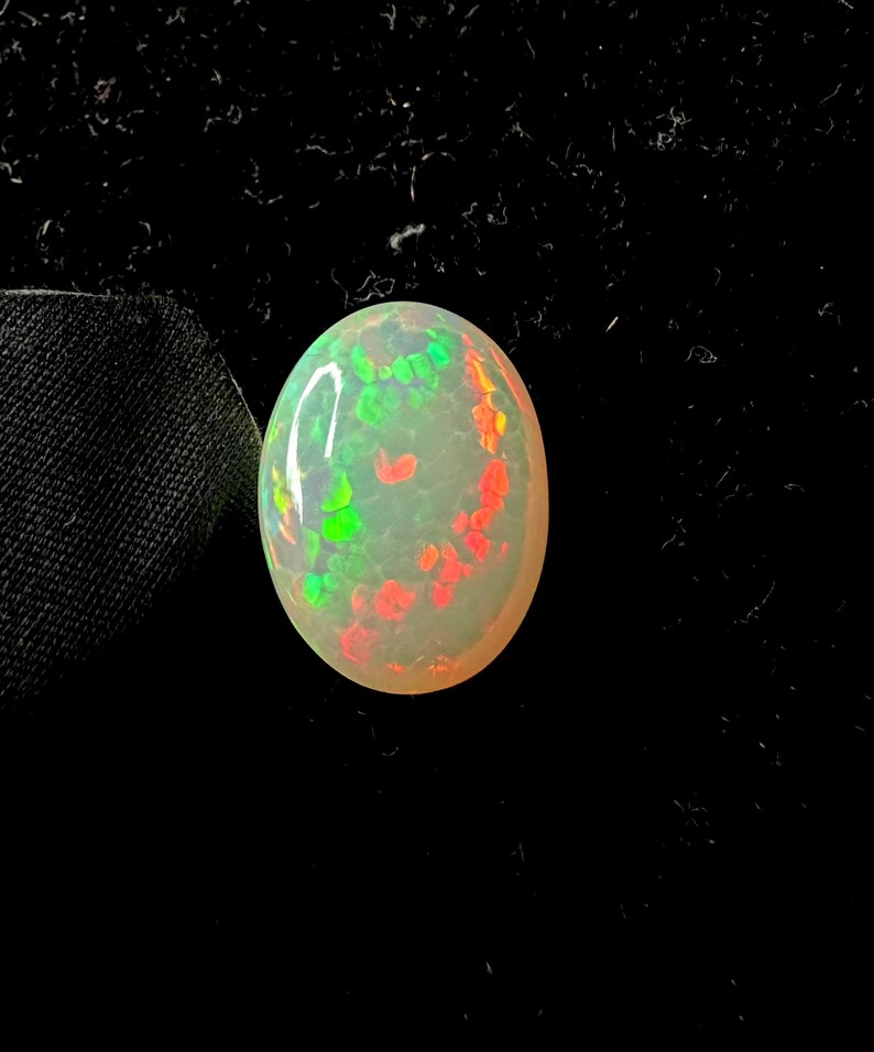 May include: An oval-shaped cabochon opal gemstone featuring a translucent, milky white base with vibrant, scattered flashes of neon green and bright orange color play across its smooth, polished, and rounded surface against a dark background.