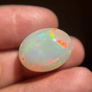 May include: An oval-shaped cabochon opal gemstone featuring a translucent, milky white base color. The stone displays a vibrant play-of-color with distinct flashes of bright red, neon green, and electric blue light scattered across its smooth surface.