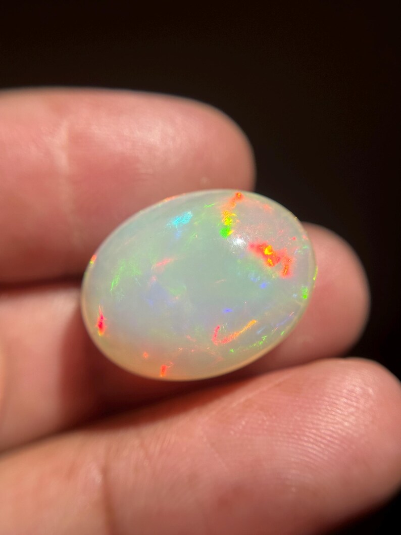 May include: An oval-shaped cabochon opal gemstone featuring a translucent, milky white base color. The stone displays a vibrant play-of-color with distinct flashes of bright red, neon green, and electric blue light scattered across its smooth surface.