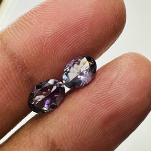 May include: Two oval-cut gemstones with a deep purple and violet hue. The stones feature a faceted surface that reflects light, creating a sparkling appearance. Each gemstone has a polished finish and a translucent quality, showcasing a rich color.