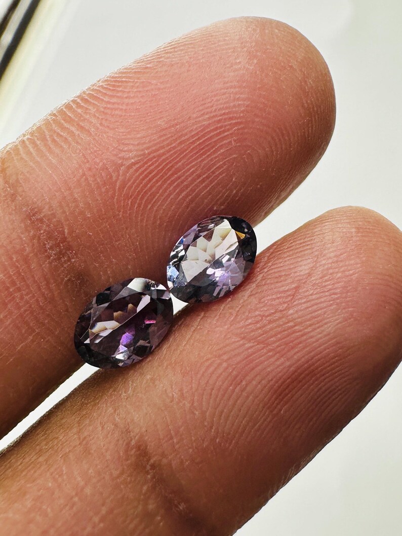 May include: Two oval-cut gemstones with a deep purple and violet hue. The stones feature a faceted surface that reflects light, creating a sparkling appearance. Each gemstone has a polished finish and a translucent quality, showcasing a rich color.
