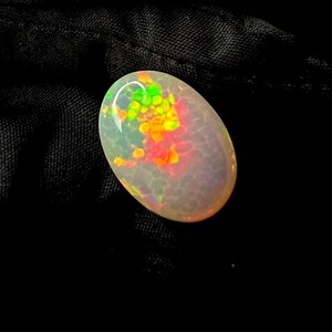 May include: An oval-shaped cabochon opal gemstone featuring a translucent, milky white base with a distinct honeycomb pattern. The stone displays vibrant flashes of neon green, bright yellow, and intense orange play-of-color across its surface.
