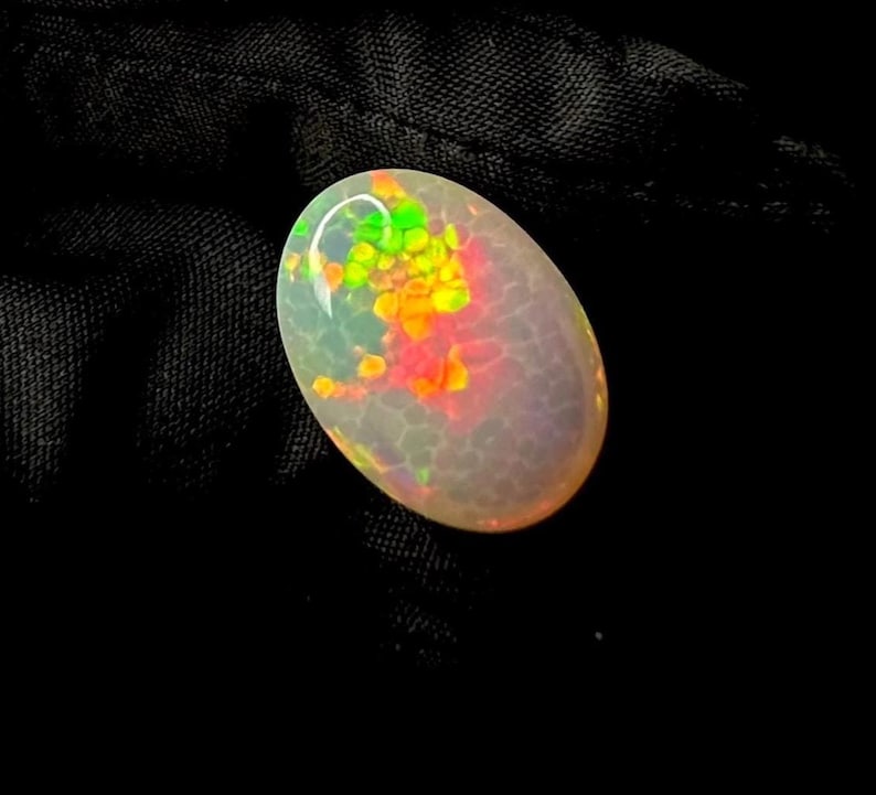 May include: An oval-shaped cabochon opal gemstone featuring a translucent, milky white base with a distinct honeycomb pattern. The stone displays vibrant flashes of neon green, bright yellow, and intense orange play-of-color across its surface.