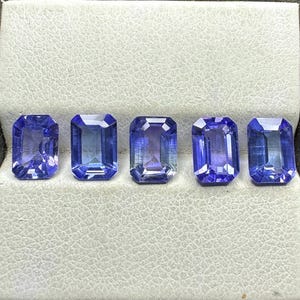 以下が含まれることがあります： 白い表面に並んだ、長方形のエメラルドカットの青い宝石が5つあります。