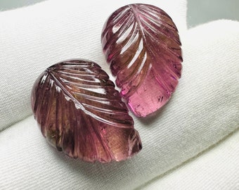 May include: Two carved gemstone leaves in shades of pink and brown. The leaves are detailed with carved veins and have a polished, translucent appearance. The gemstones are set against a white background.