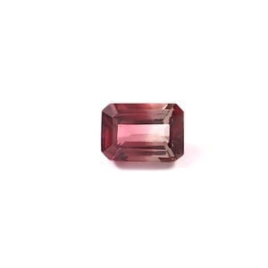 May include: A rectangular-cut gemstone with a gradient of colors, transitioning from a deep burgundy at the edges to a lighter pinkish hue in the center. The gem is set against a plain white background, highlighting its facets and color variations.