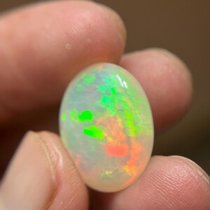 May include: An oval-shaped cabochon opal gemstone featuring a translucent white base with vibrant flashes of neon green and orange play-of-color throughout the stone. The gemstone has a smooth, polished surface and a rounded, symmetrical profile.