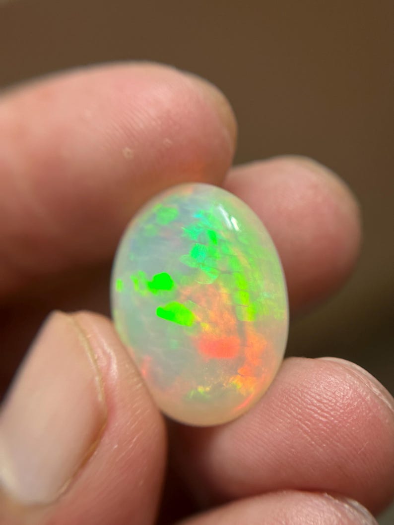 May include: An oval-shaped cabochon opal gemstone featuring a translucent white base with vibrant flashes of neon green and orange play-of-color throughout the stone. The gemstone has a smooth, polished surface and a rounded, symmetrical profile.
