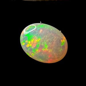 May include: An oval-shaped cabochon gemstone with a translucent, milky white base. The stone displays a vibrant play-of-color, featuring scattered flashes of neon green, bright orange, and yellow hues across its smooth, polished surface.