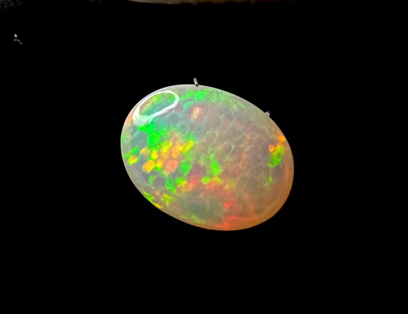 May include: An oval-shaped cabochon gemstone with a translucent, milky white base. The stone displays a vibrant play-of-color, featuring scattered flashes of neon green, bright orange, and yellow hues across its smooth, polished surface.