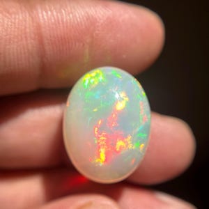 May include: An oval-shaped cabochon opal gemstone featuring a translucent white base with vibrant flashes of red, orange, yellow, and green play-of-color throughout the stone. The gemstone has a smooth, polished surface and a rounded dome shape.