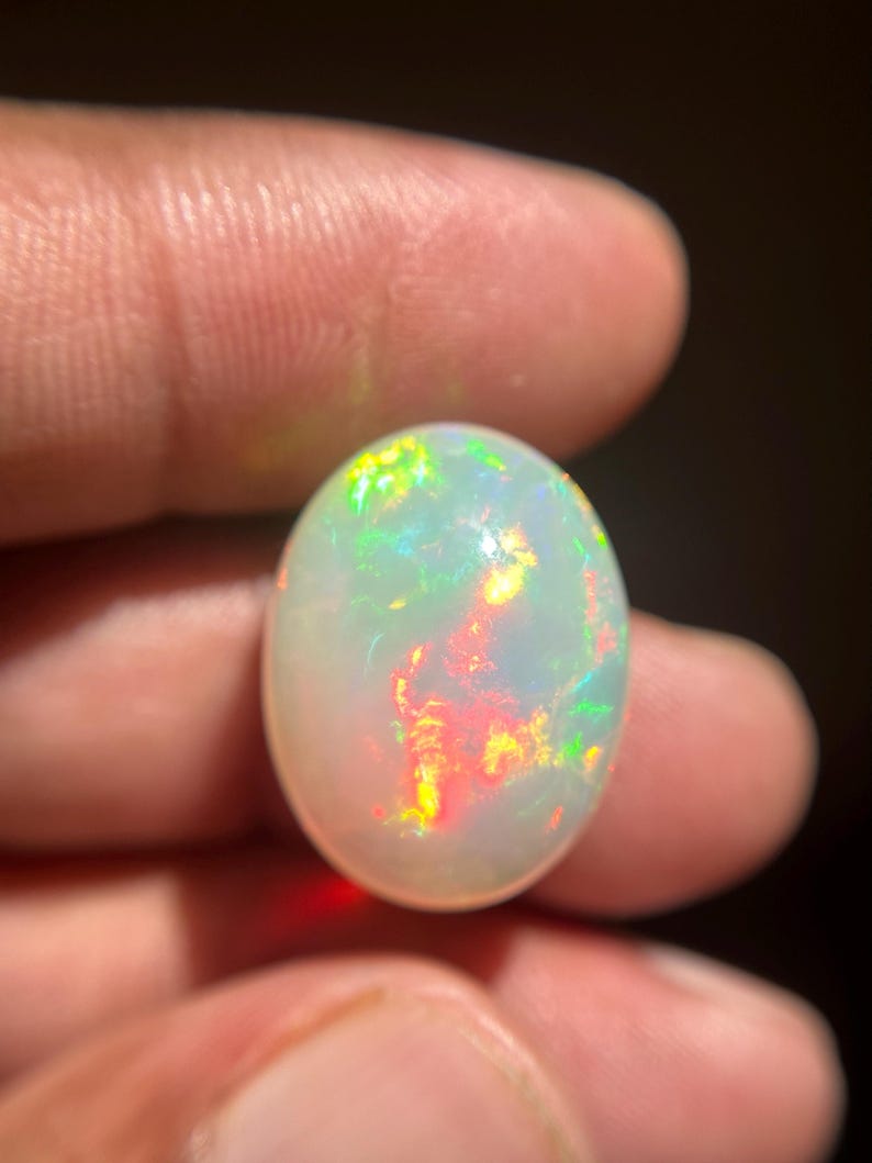 May include: An oval-shaped cabochon opal gemstone featuring a translucent white base with vibrant flashes of red, orange, yellow, and green play-of-color throughout the stone. The gemstone has a smooth, polished surface and a rounded dome shape.