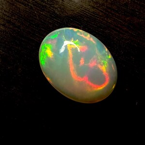 May include: An oval-shaped cabochon opal gemstone featuring a translucent white base with vibrant flashes of red, orange, and green play-of-color. The stone has a smooth, polished surface that reflects light, highlighting the internal color patterns.