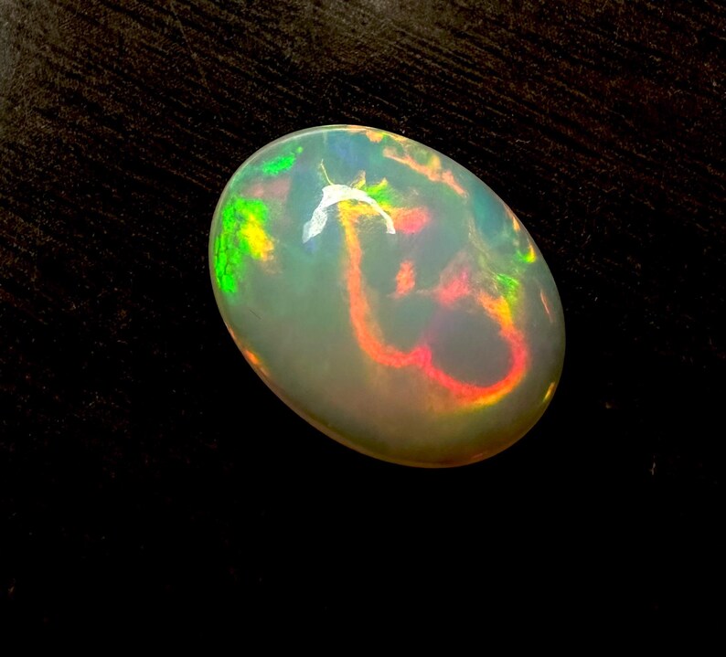 May include: An oval-shaped cabochon opal gemstone featuring a translucent white base with vibrant flashes of red, orange, and green play-of-color. The stone has a smooth, polished surface that reflects light, highlighting the internal color patterns.